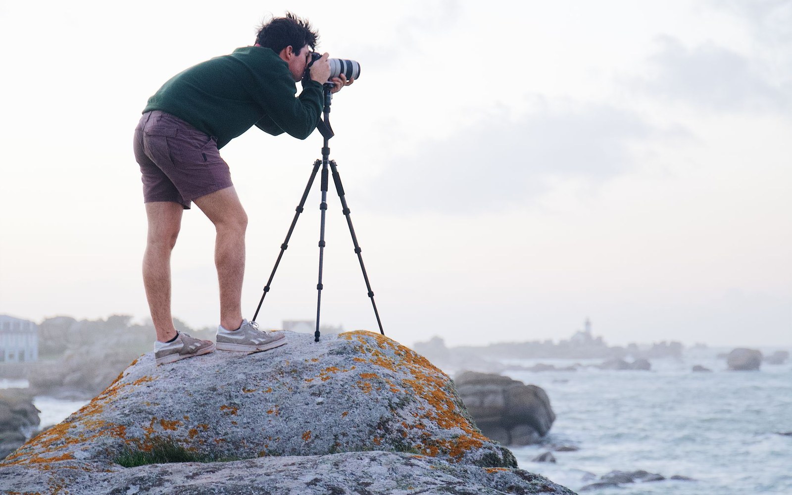 A cinematographer at the waterline with a 70–200mm lens on a monopod, shooting toward an incoming set