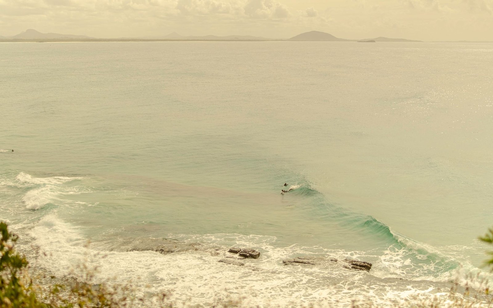 A lone surfer rides a breaking wave off a rocky, hazy coastline