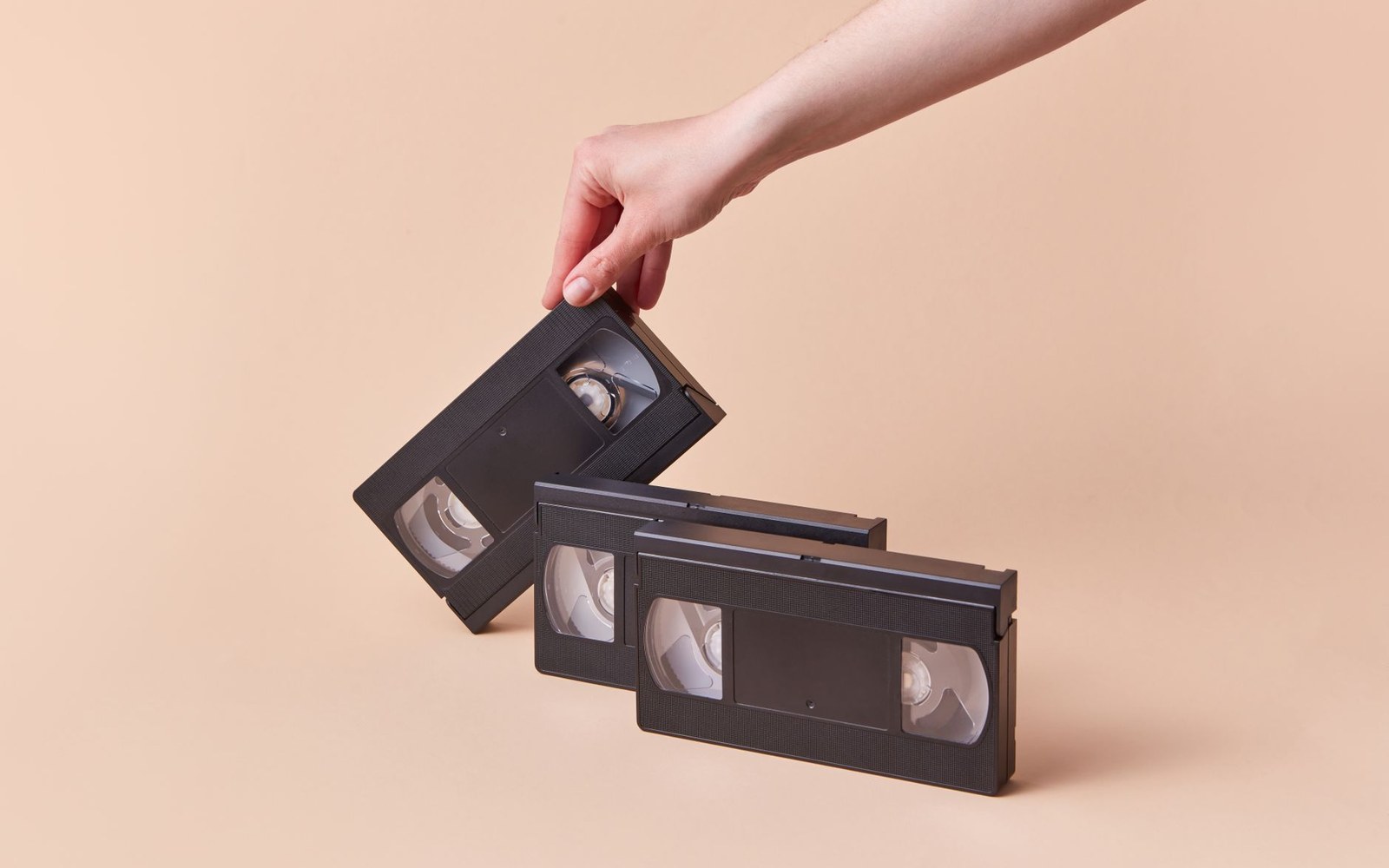 A hand picks up a VHS tape from a stack of three against a beige background